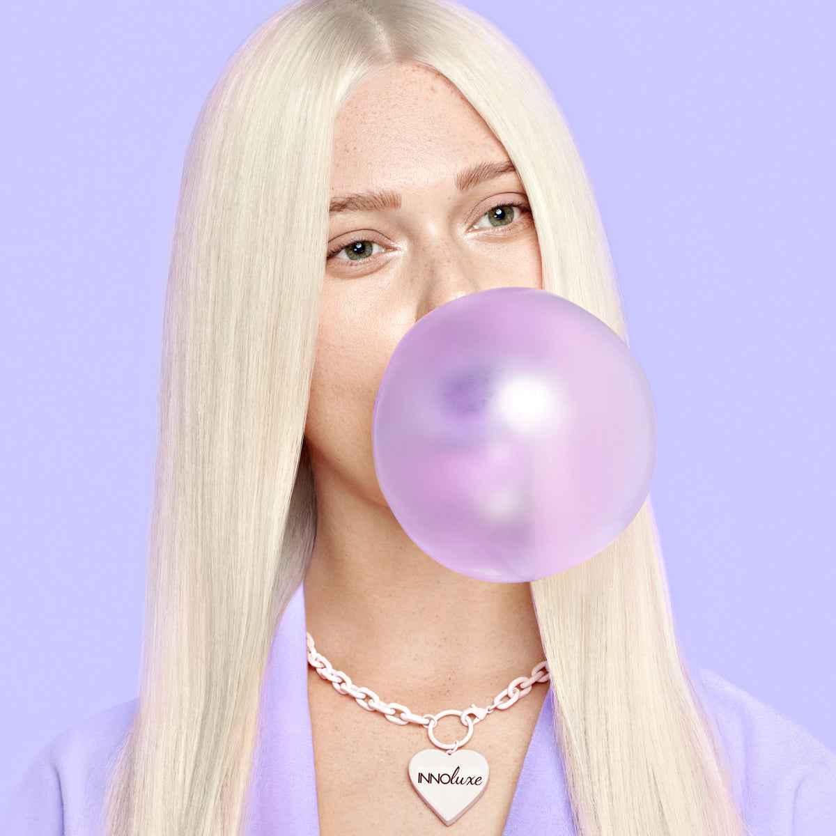 Woman with cool platinum blonde toned hair against a lavender background, blowing a violet bubble — hair toner guide